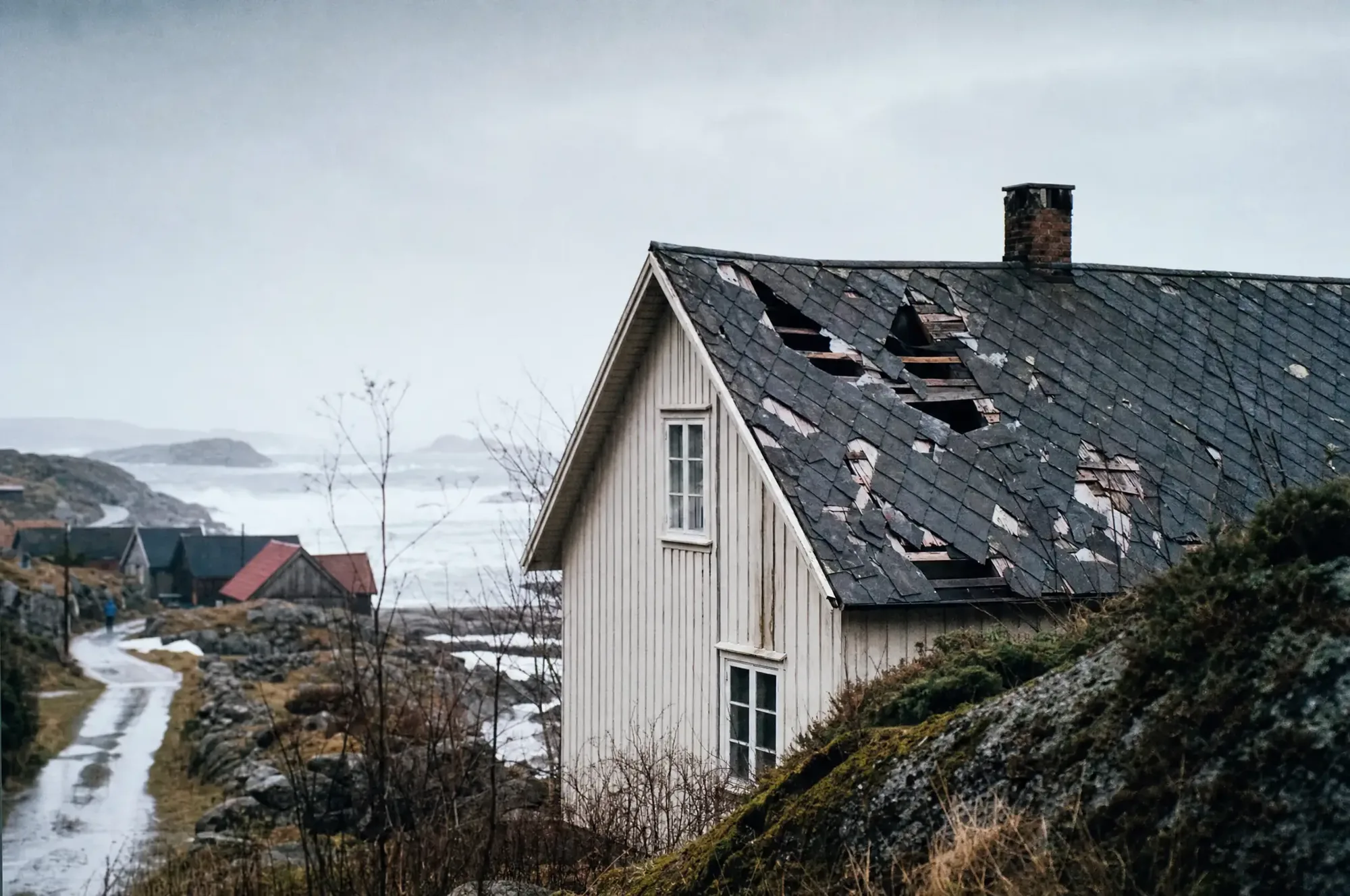 Skadet takstein på et trehus i kystlandskap etter storm, med vått underlag