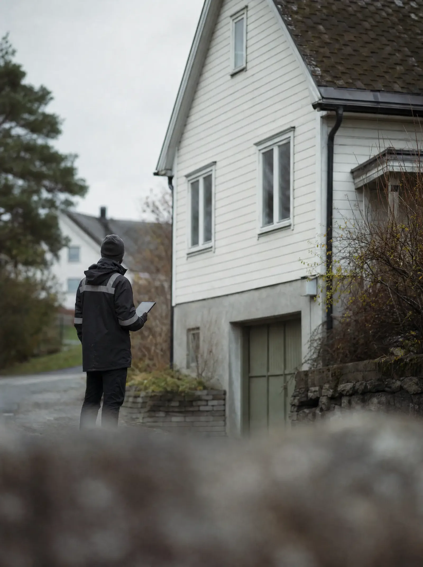 Takstmann med nettbrett ved en trekledd bolig i et boligfelt i overskyet vær