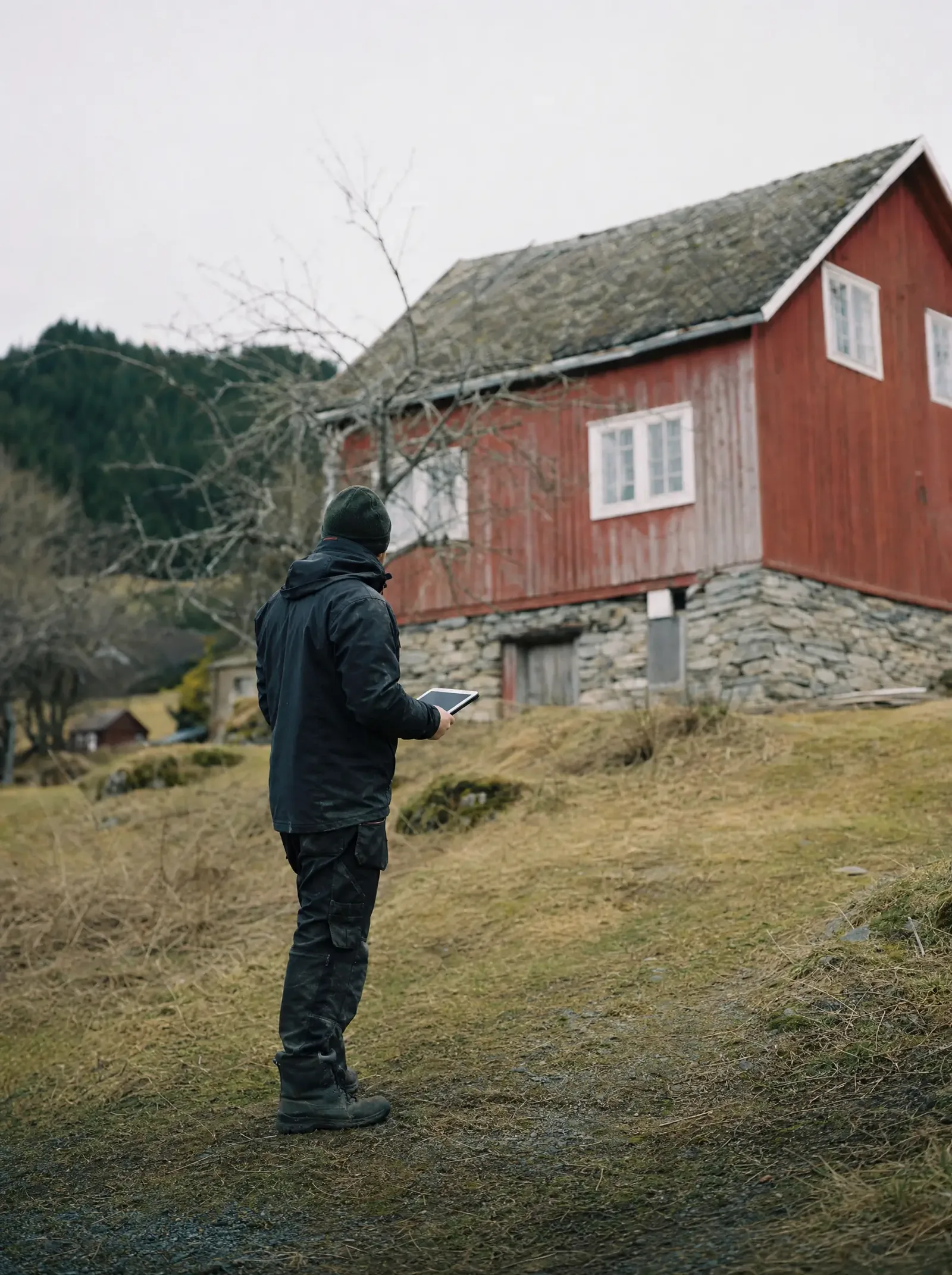 Takstmann i mørk arbeidsjakke ved et eldre rødmalt våningshus i landlige omgivelser, fotografert på avstand